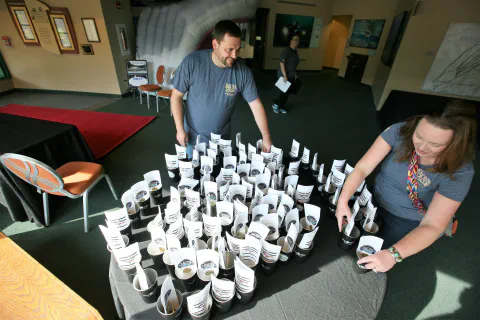 Two people arrange numerous small cups with paper tags on a table in a spacious indoor setting.