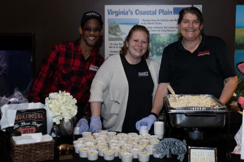 Three people smile while serving food at an event, with a sign reading Virginia's Coastal Plain in the background.