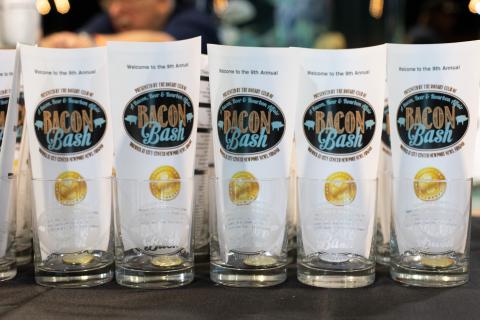 Row of clear glasses with "Bacon Bash" printed on them, each wrapped in a branded sleeve.