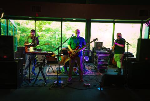 Four musicians perform on stage with guitars, keyboards, and drums, bathed in colorful stage lights against a green backdrop.