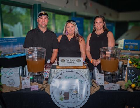 Three smiling staff members stand behind a booth promoting their mobile saloon, featuring large pitchers of lemonade.