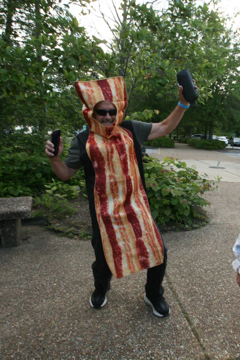 Person dressed as bacon, holding controllers, smiling outdoors with trees and pavement in background